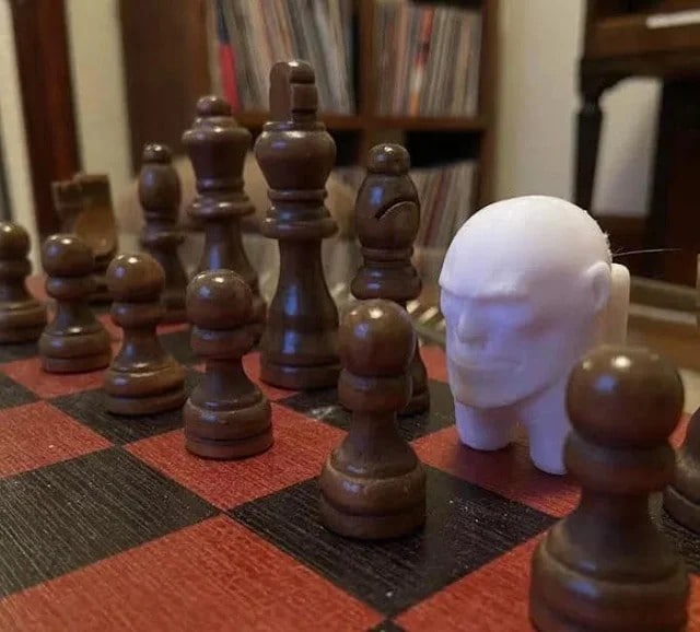 Among Us голова на шахматной доске 3D-печатная фигурка Among Us, которая выглядит как скульптура человеческой головы, стоит на шахматной доске среди шахматных фигур.