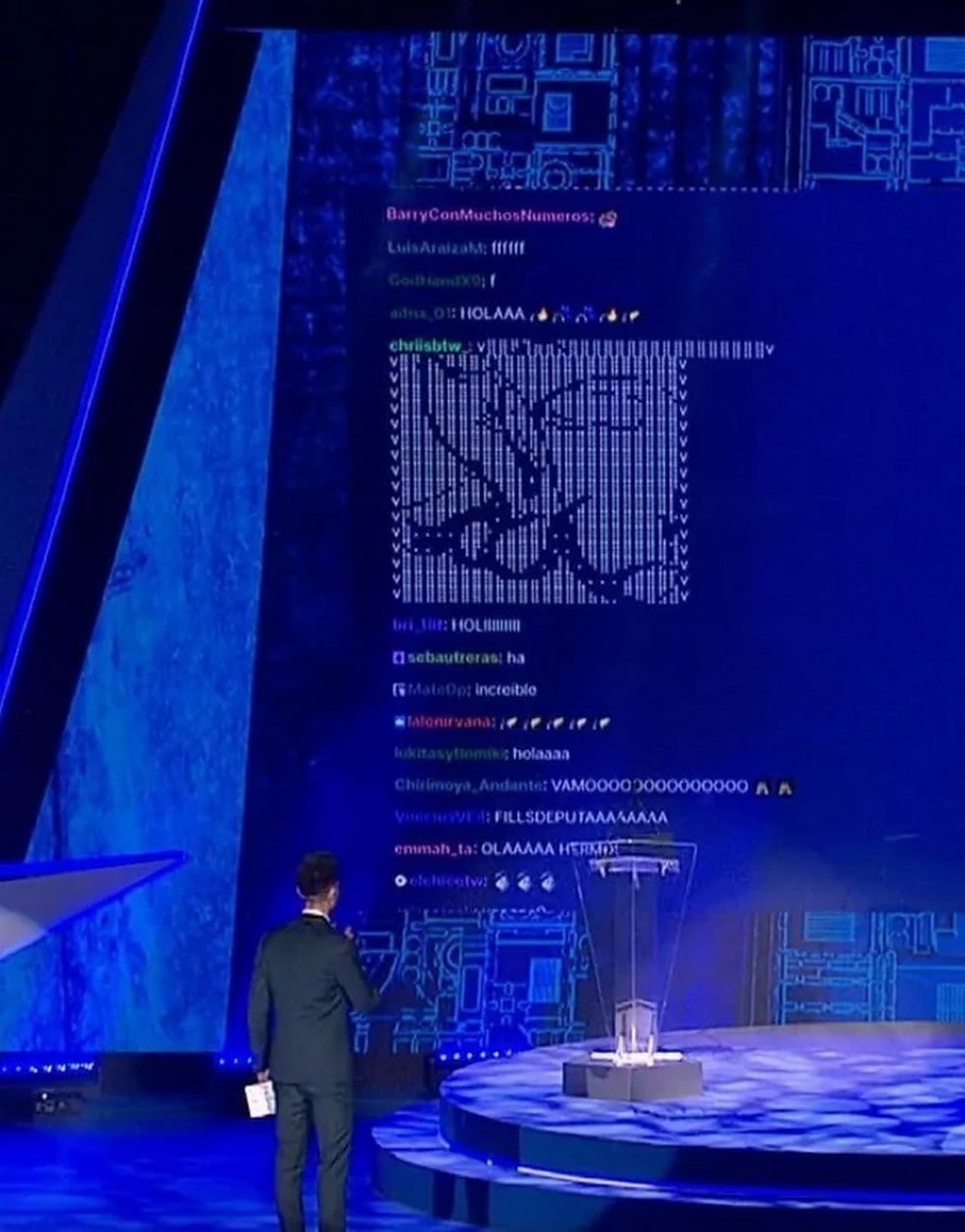 ASCII-арт в чате трансляции Ведущий на сцене стоит спиной к большому экрану, на котором отображаются сообщения из чата, включая ASCII-арт.