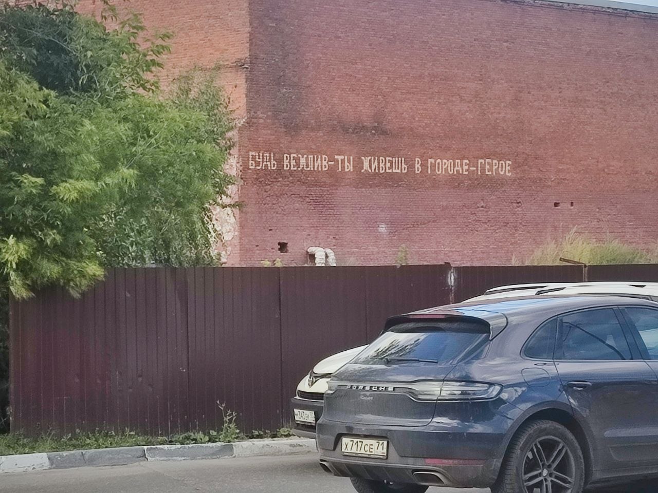 Надпись на кирпичной стене: «Будь вежлив — ты живёшь в городе-герое», снятая на фоне городского забора и автомобилей.