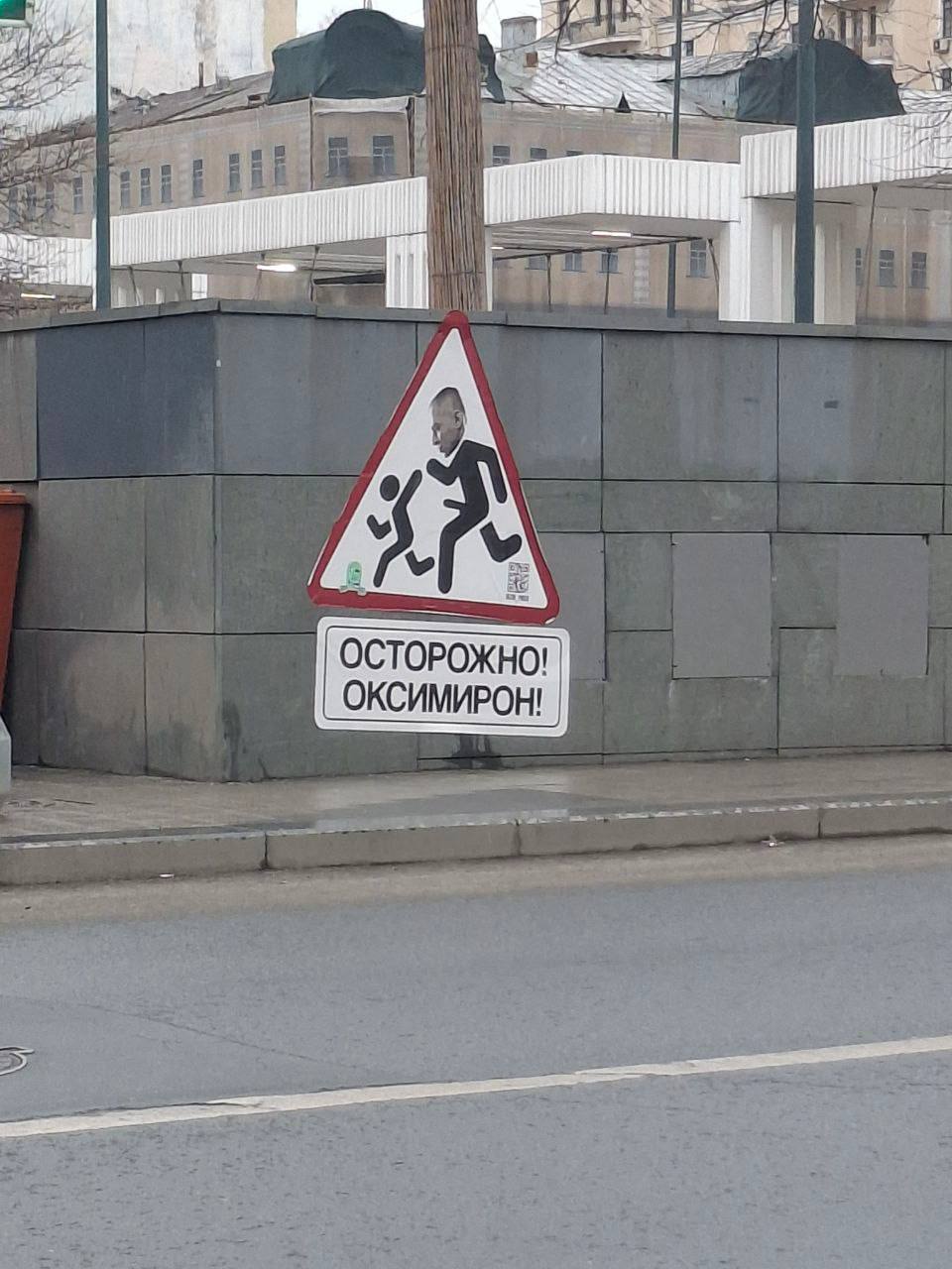 Осторожно, Оксимирон Дорожный знак «Осторожно, дети» с лицом Оксимирона, установленный на бетонном ограждении рядом с проезжей частью. Под знаком находится табличка с надписью «Осторожно! Оксимирон!»