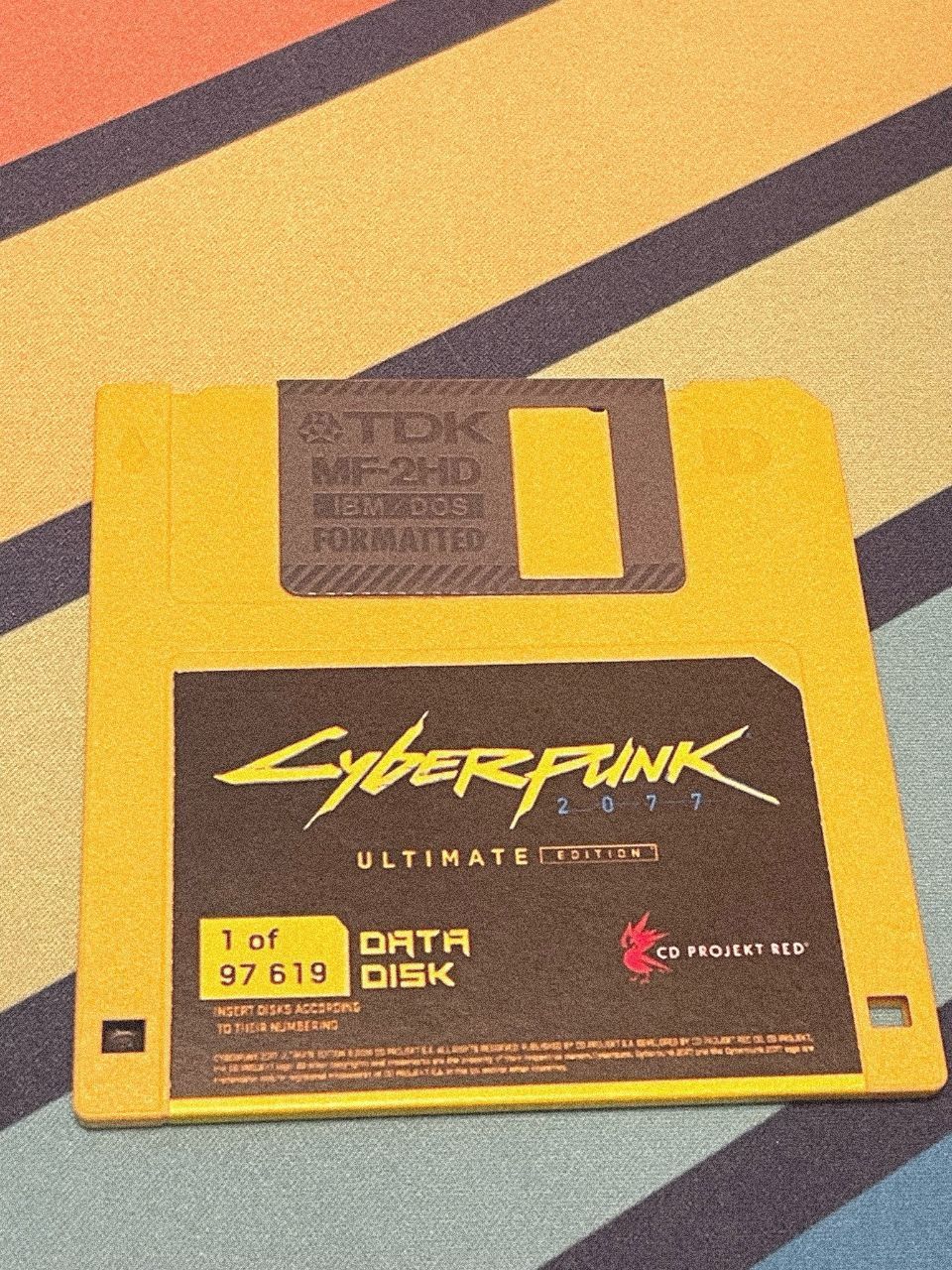 Cyberpunk 2077 на дискете Желтая дискета с надписью Cyberpunk 2077 Ultimate Edition на полосатом фоне