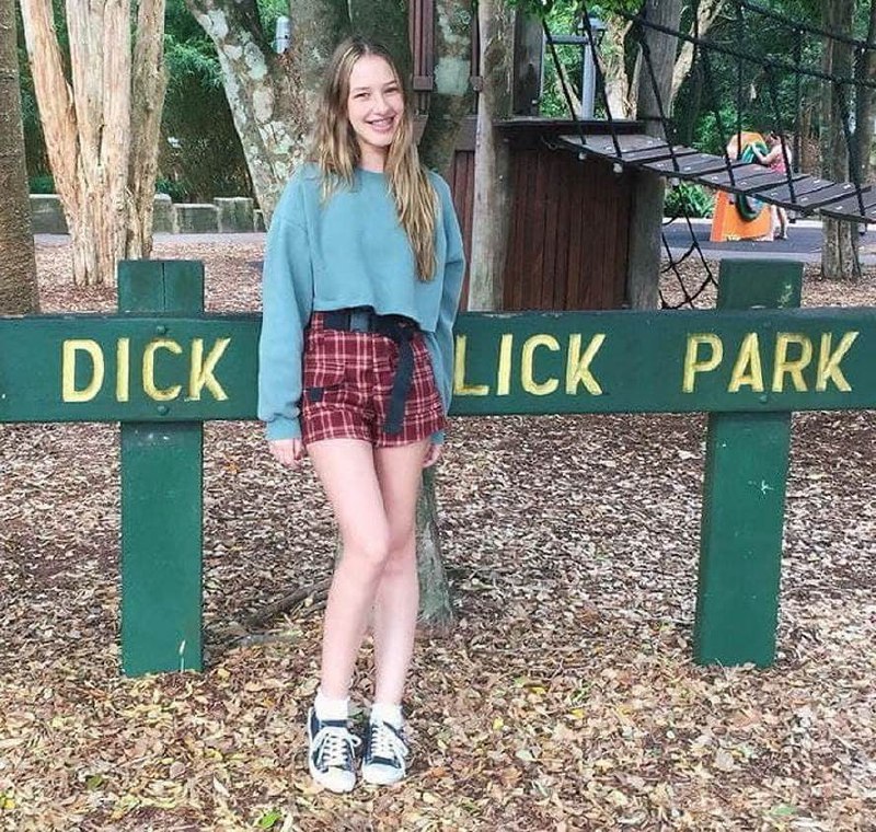Девушка у таблички Dick Lick Park Мем с девушкой, стоящей у зелёной таблички с надписью Dick Lick Park, на фоне деревьев и элементов детской площадки