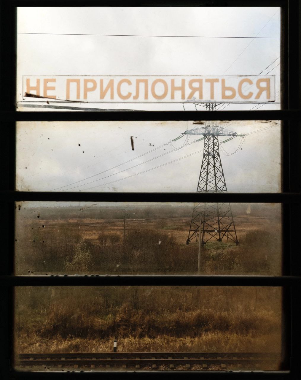 Вид из окна поезда на поле и линию электропередачи с необычным указателем «Не приспосабляться».