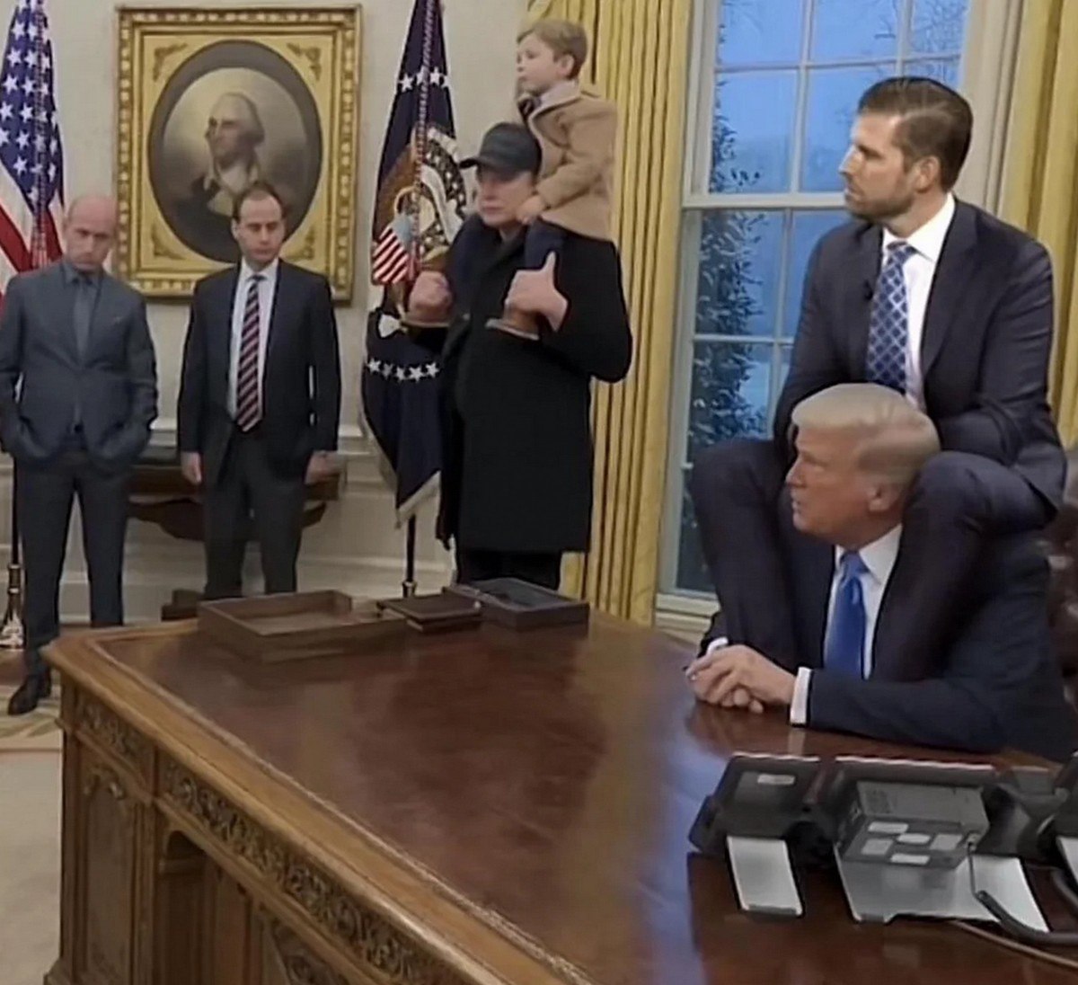 Дональд Трамп, Илон Маск и их сыновья Дональд Трамп сидит за столом, а на его плечах сидит его сын Дональд Трамп-младший, рядом стоит Илон Маск с ребенком на плечах в Овальном кабинете.