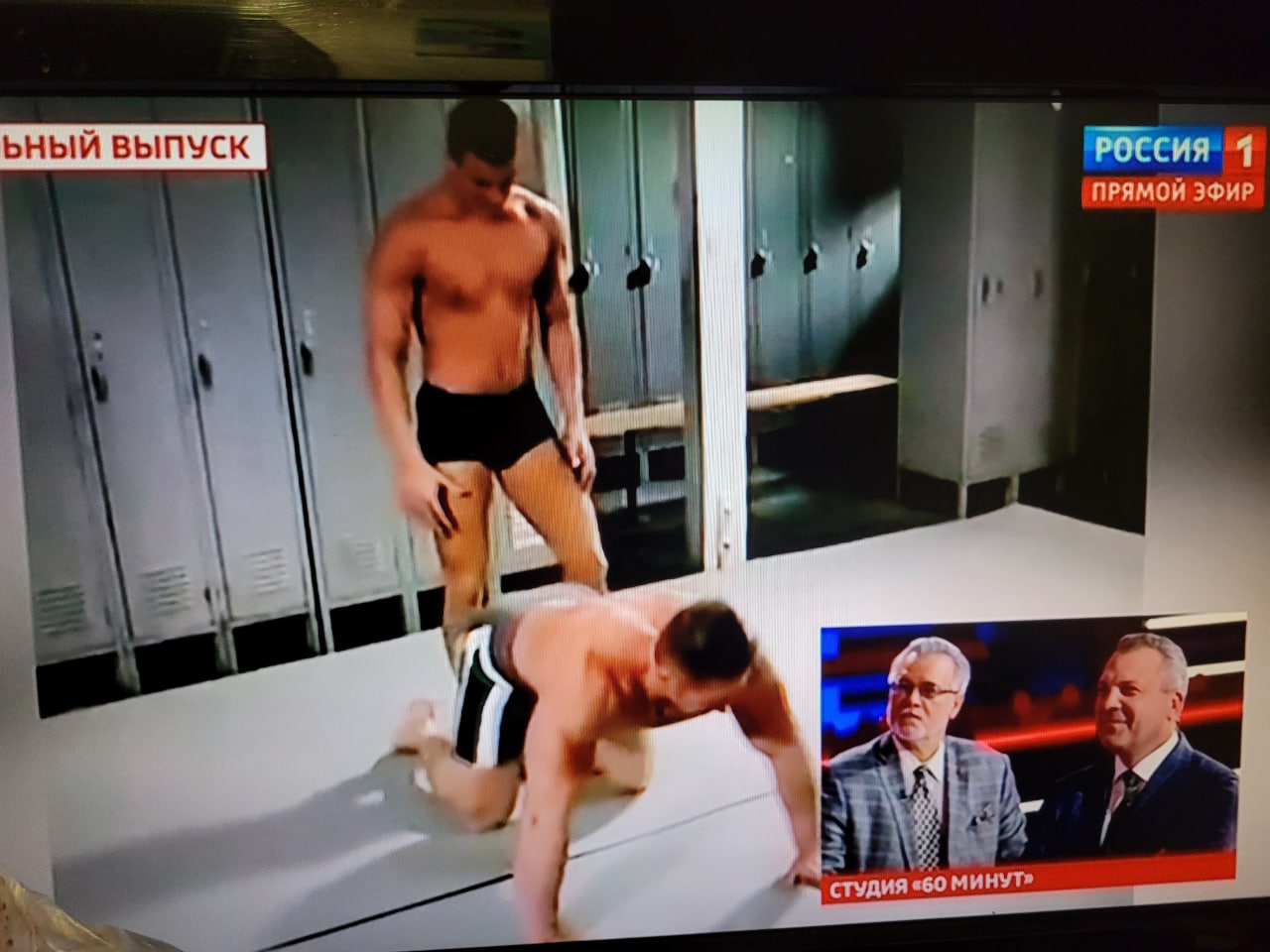 Двое мужчин в раздевалке на фоне ведущих 60 минут Двое мужчин в раздевалке в шортах, один стоит, другой на четвереньках, на фоне телеведущих "60 минут" и логотипов канала "Россия 1"