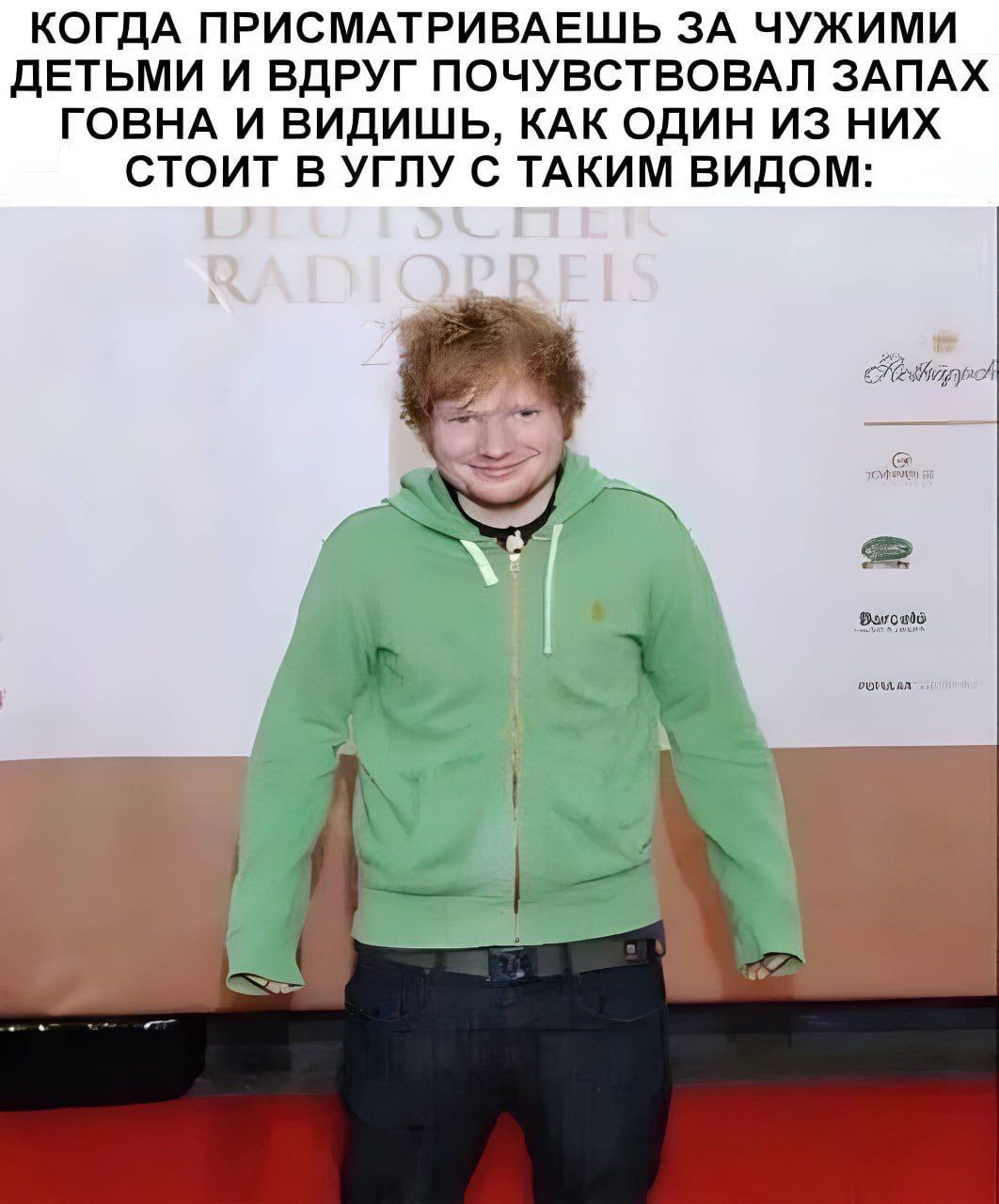 Эд Ширан улыбается и что-то скрывает Эд Ширан в зеленой худи, стоит и хитро улыбается на фоне бежевой стены с надписями, вероятно, скрывая что-то.