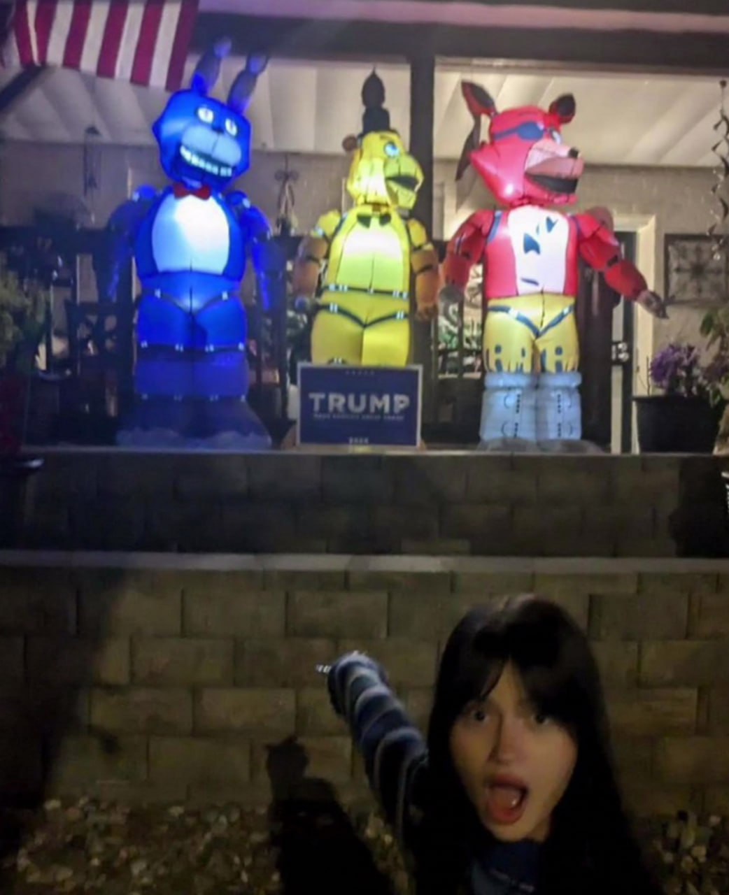 FNAF и табличка Трампа на крыльце Надувные фигуры персонажей из Five Nights at Freddy's (FNAF) на крыльце дома, между которыми стоит табличка "Трамп", а девушка указывает на них пальцем.