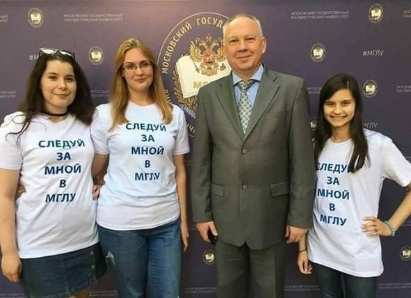 Три девушки в футболках с надписью «Следуй за мной в МГПУ» фотографируются с мужчиной в костюме на фоне эмблемы университета.