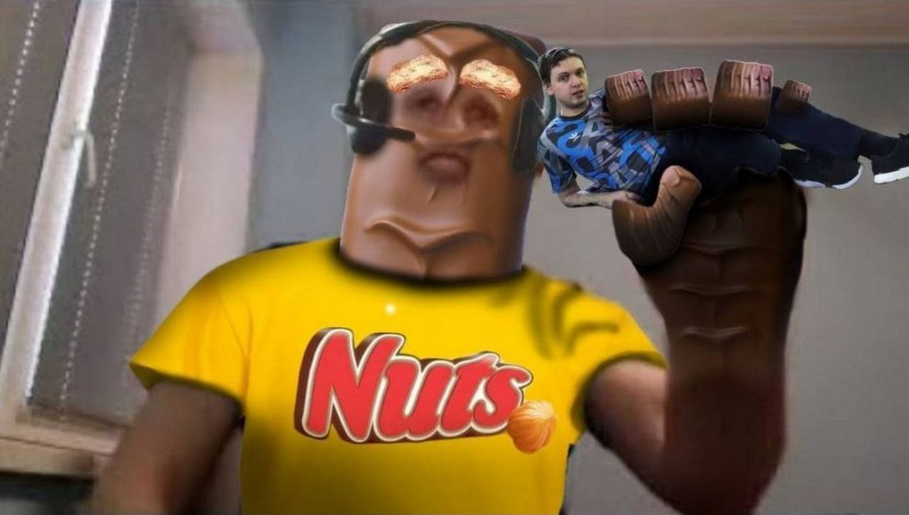 Голова Nuts Персонаж с головой батончика Nuts и телом человека в желтой футболке с логотипом Nuts, держащий на руке другого человека