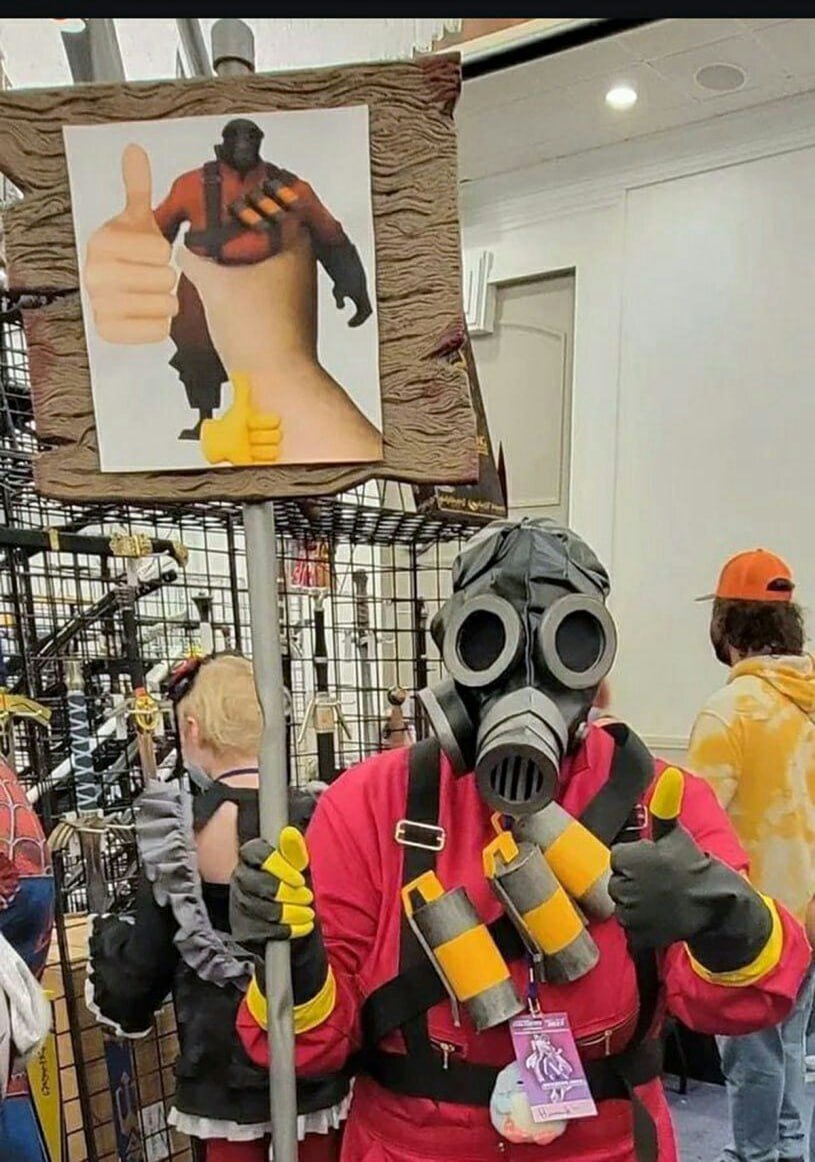 Хэви обнимает большой палец вверх Косплеер Пиро из Team Fortress 2 держит табличку с Хэви и рукой, показывающей большой палец вверх.