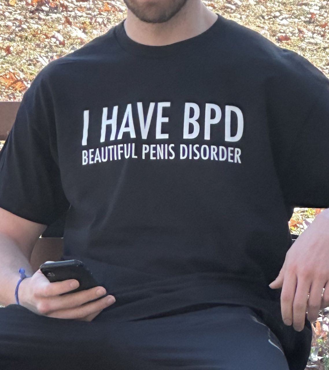 Футболка с надписью I have BPD beautiful penis disorder Мужчина в черной футболке с надписью "I HAVE BPD BEAUTIFUL PENIS DISORDER" сидит на скамейке и смотрит в телефон.