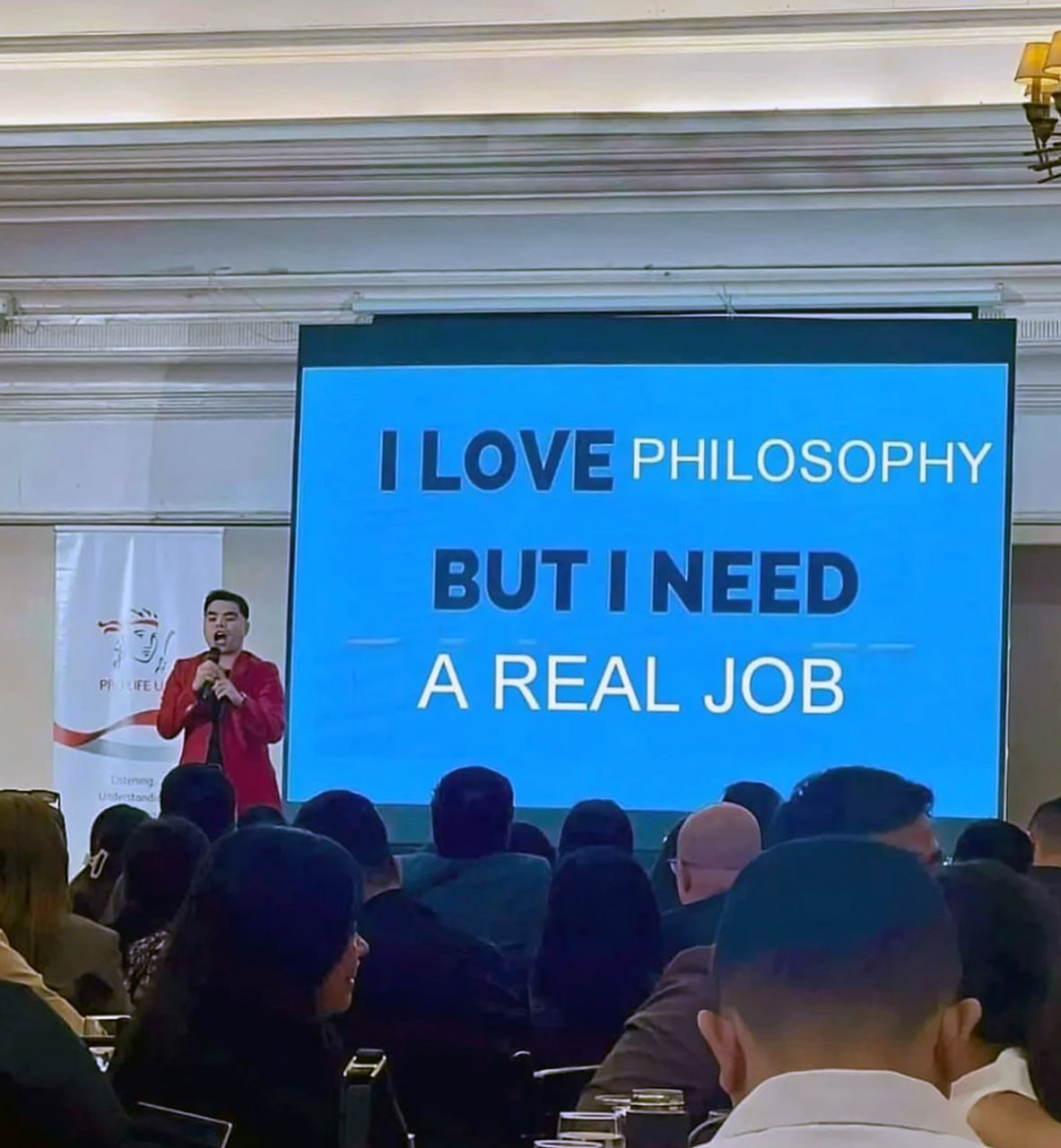 I love philosophy but I need a real job Мужчина выступает перед аудиторией, а на экране позади него надпись «Я люблю философию, но мне нужна настоящая работа».
