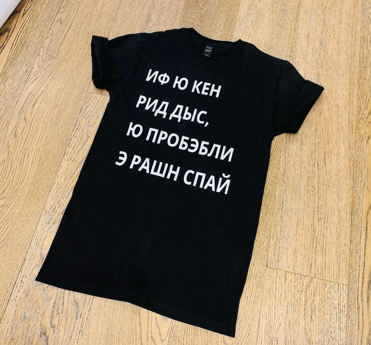 Черная футболка с надписью "ИФ Ю КЕН РИД ДЫС, Ю ПРОБЭБЛИ Э РАШН СПАЙ" на деревянном полу