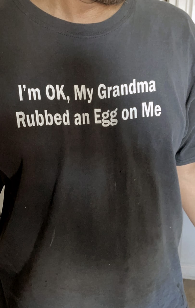 I'm OK, My Grandma Rubbed an Egg on Me Мужчина в черной футболке с надписью «I’m OK, My Grandma Rubbed an Egg on Me» крупным планом