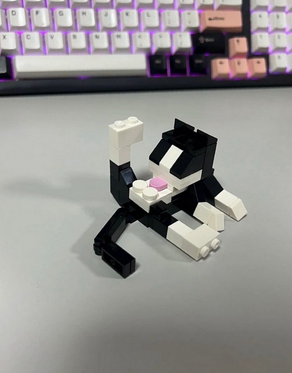 Кот из лего лежит Собранный из конструктора Lego кот в черно-белых тонах лежит на столе на фоне белой клавиатуры с подсветкой