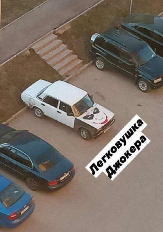Старый белый автомобиль с рисунком лица Джокера на капоте и забавной подписью.