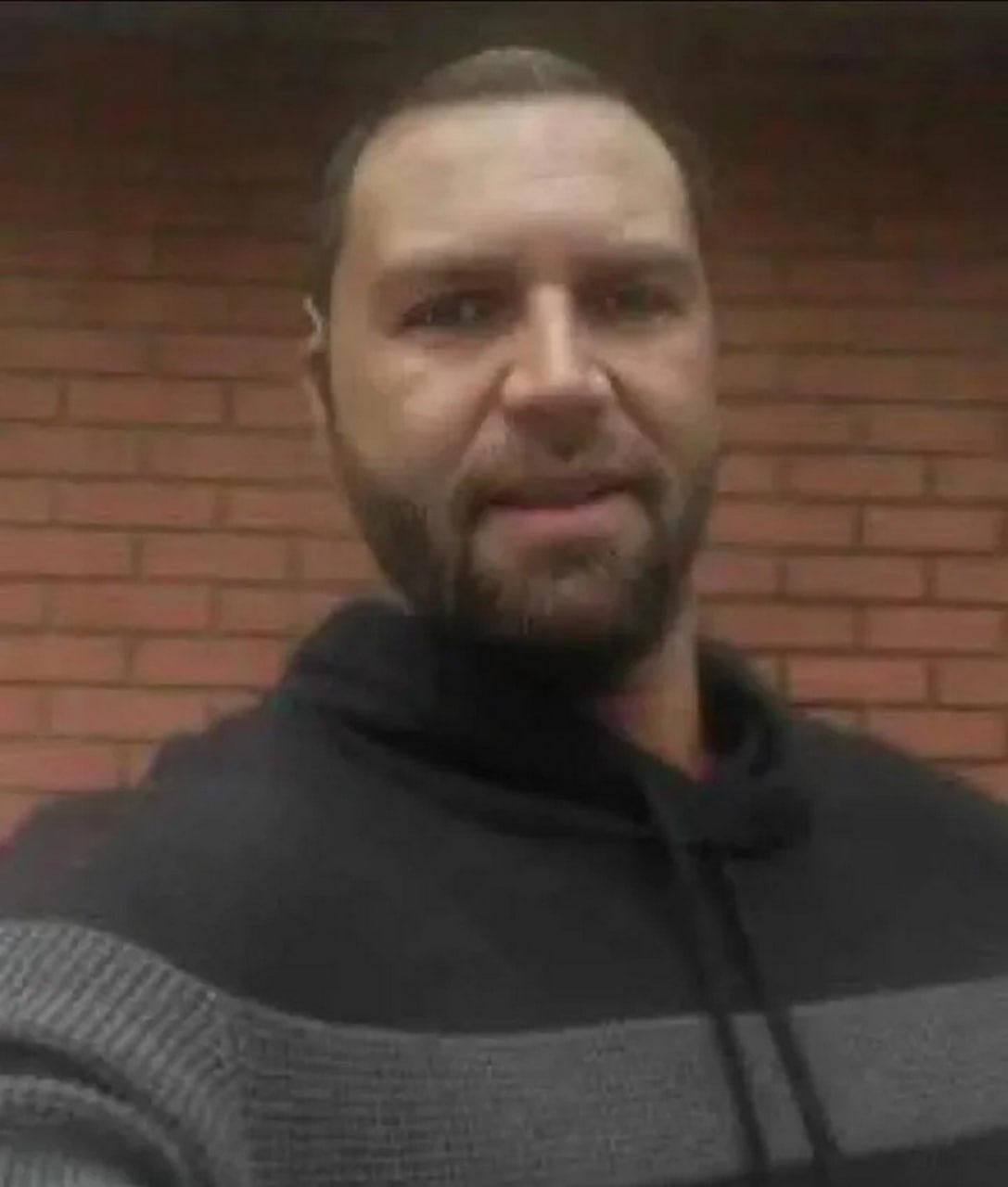 Мужчина в свитере с бородой на фоне кирпичной стены Бородатый мужчина в свитере с капюшоном, смотрящий прямо в камеру на фоне кирпичной стены.