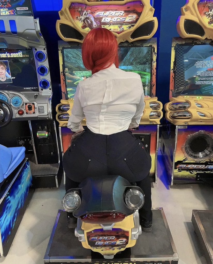 Мужчина с рыжими волосами играет в Super Bikes 2 Мужчина с рыжими волосами сидит на игровом автомате Super Bikes 2 в аркадном зале