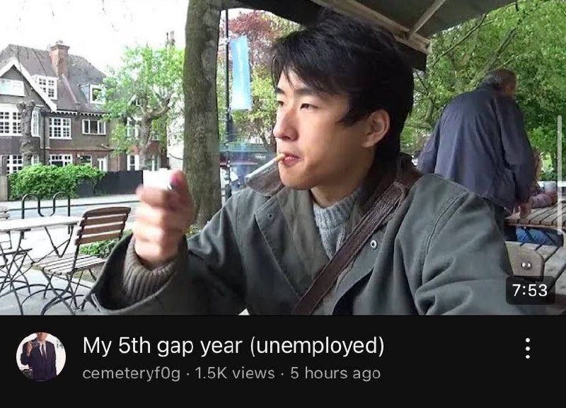 My 5th gap year (unemployed) Мужчина азиатской внешности прикуривает сигарету на улице. Он сидит за столом и смотрит в сторону. Возможно, это кадр из видеоблога.