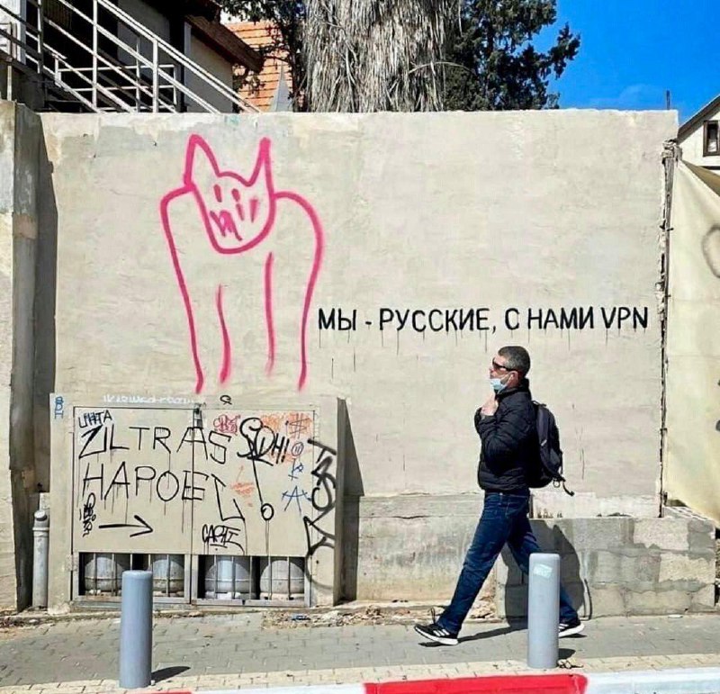 Мы - русские, с нами VPN Граффити «Мы - русские, с нами VPN» на стене с изображением розового кота, мимо которой проходит мужчина в маске и с рюкзаком.