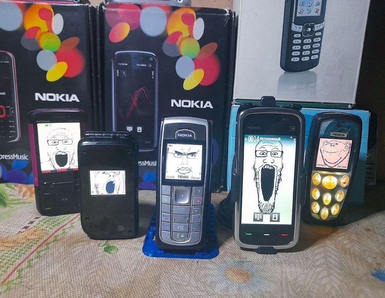 Nokia Soyjack wojak phones Куча старых телефонов Nokia с экранами, на которых изображены вариации мема «Вояк» (wojak), включая Soyjack и Yes Chad. Телефоны стоят на столе, а за ними виднеются коробки от этих же телефонов.