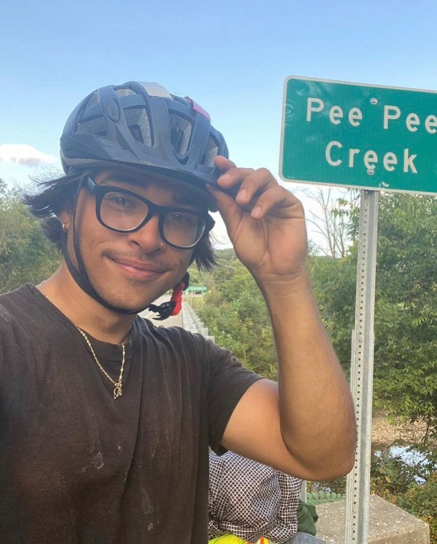 Pee Pee Creek Мужчина в очках и шлеме для велосипеда стоит на фоне таблички с названием ручья «Pee Pee Creek».