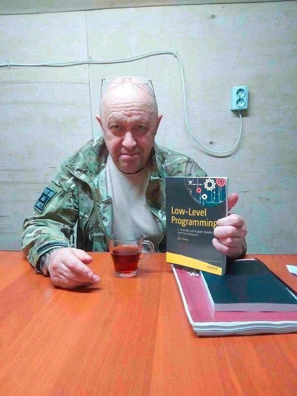 Пригожин с книгой по программированию Евгений Пригожин сидит за столом с кружкой чая и книгой "Low-Level Programming".