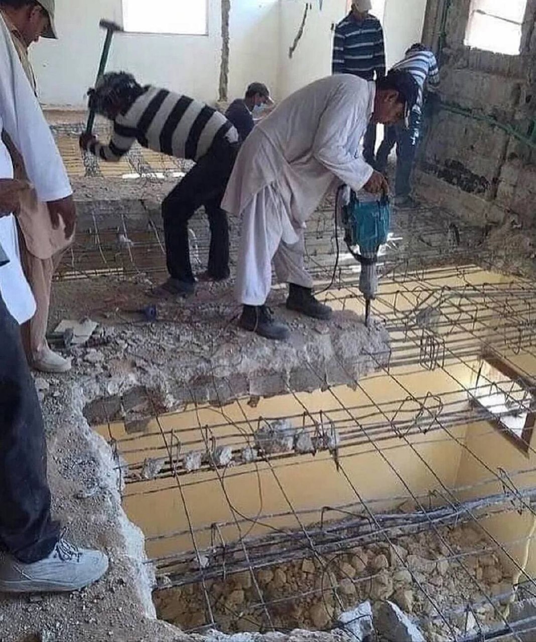 Рабочие ломают пол Рабочие используют кувалду и отбойный молоток, чтобы ломать пол в здании. Виден арматурный каркас и отверстия, ведущие на нижний этаж.