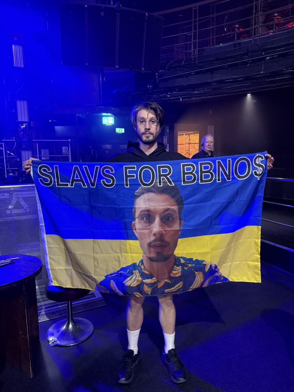 Славяне за BBNO$ На фотографии изображен мужчина, держащий украинский флаг с надписью «SLAVS FOR BBNO$» и портретом исполнителя bbno$ на нем, создавая эффект, будто он одет в футболку с бананами и его нижняя часть тела выходит из-под флага.