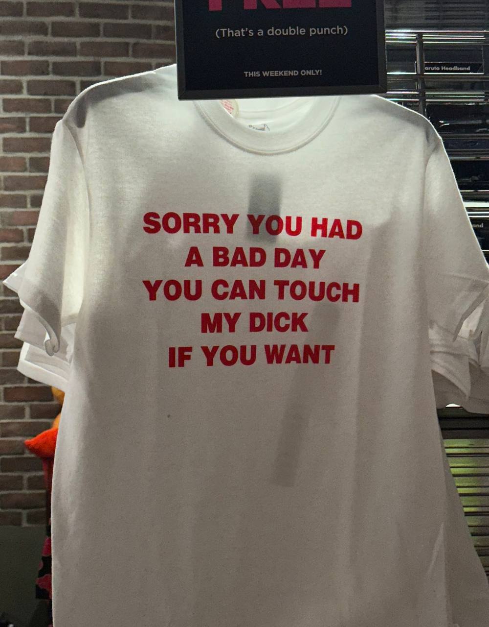 Sorry you had a bad day, you can touch my dick if you want Белая футболка с надписью «Sorry you had a bad day, you can touch my dick if you want» на вешалке в магазине. Справа видны другие футболки, а сзади стена из кирпича и какая-то вывеска.