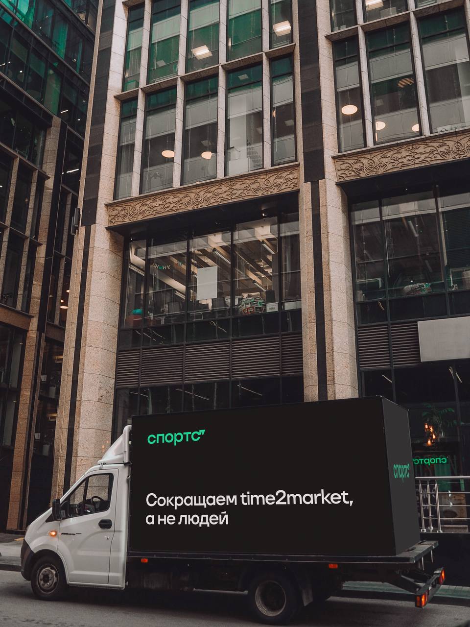 Сокращаем time2market, а не людей Фургон с надписью «Спортс. Сокращаем time2market, а не людей» на фоне современного офисного здания.