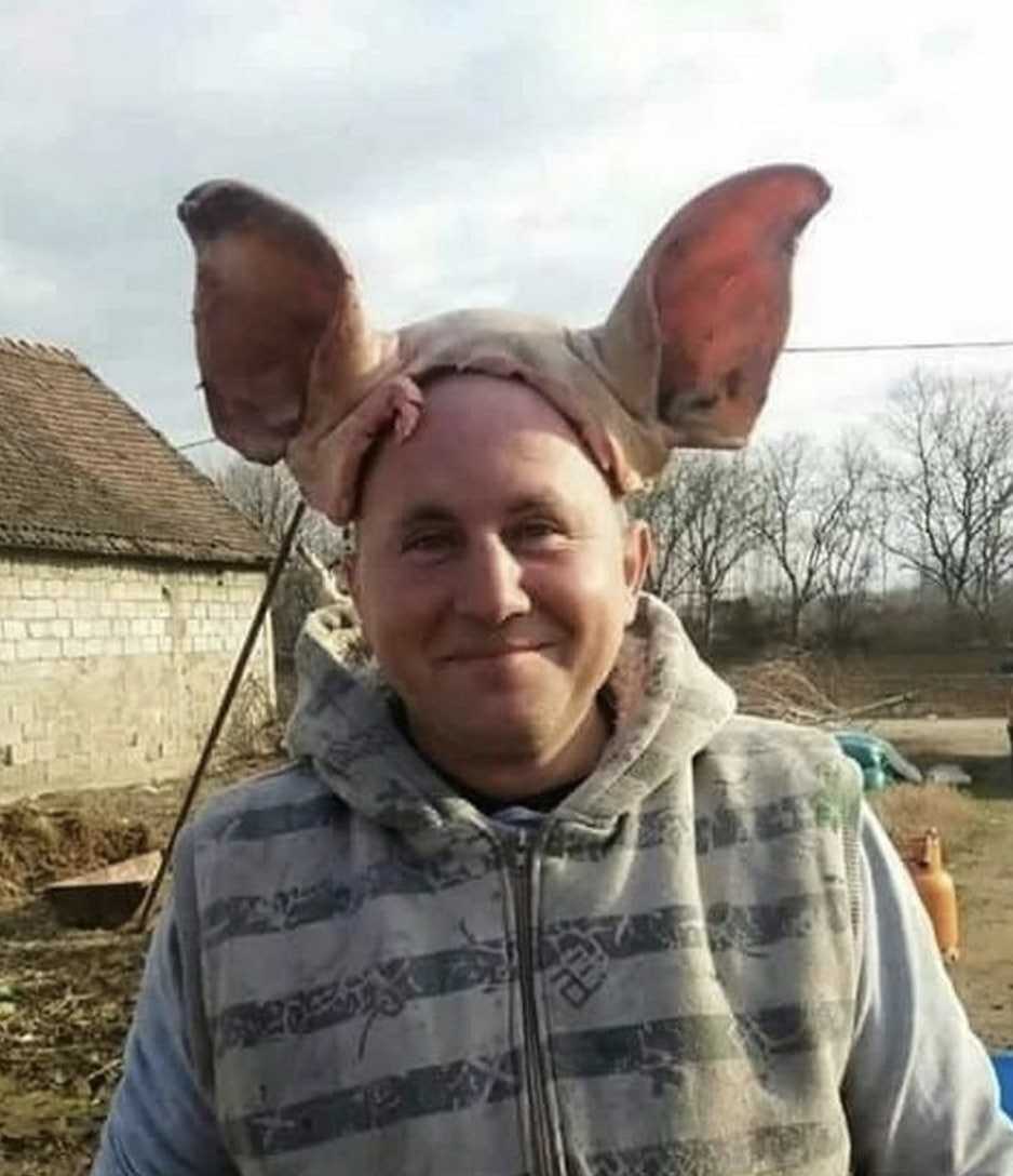 Свиные уши на голове Мужчина в жилетке с узорами на голове носит что-то похожее на свиные уши. За ним видны старый дом из кирпича и деревья без листьев.