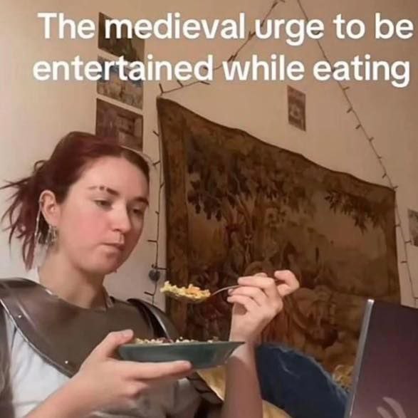 The medieval urge to be entertained while eating Девушка в доспехах ест и смотрит в ноутбук с надписью «The medieval urge to be entertained while eating»