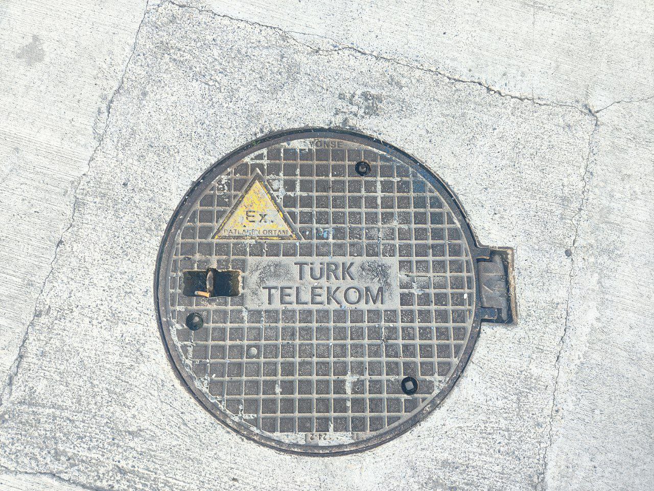 Крышка люка Turk Telekom с надписью Ex Крышка люка с надписью "Turk Telekom" и знаком "Ex" на сером асфальте