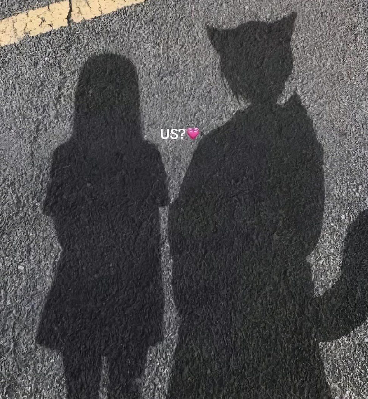 Тени двух людей на асфальте с надписью «US?» и розовым сердечком, похожие на милую пару.