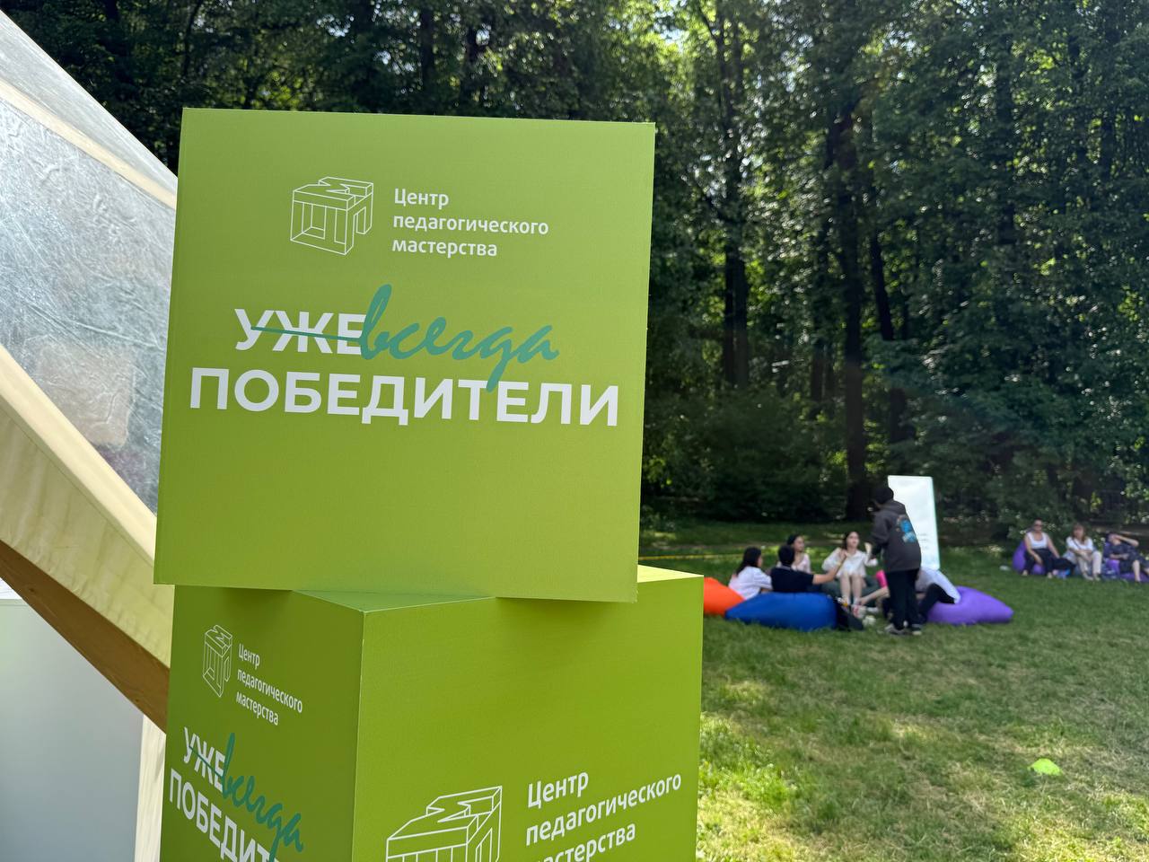 Уже всегда победители Яркие зеленые стенды с надписью "Уже всегда победители" и логотипом "Центр педагогического мастерства" на фоне людей, сидящих на пуфиках в парке.