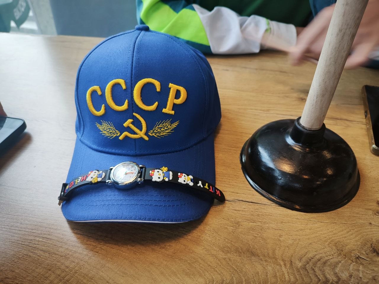 Синяя кепка с символикой СССР, часами и ремешком Hello Kitty лежит рядом с вантузом на деревянном столе.