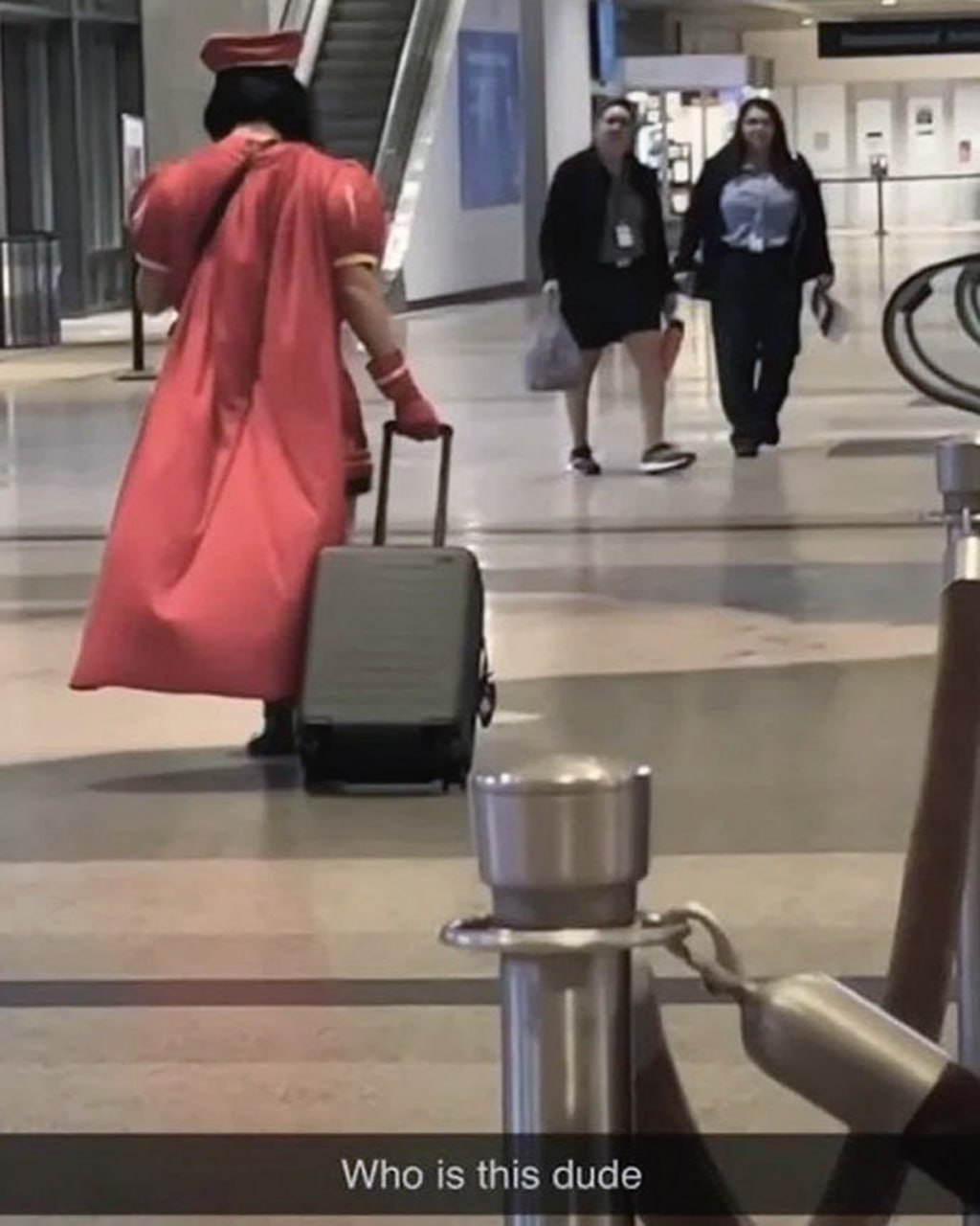 Who is this dude Человек в красной мантии в аэропорту тянет за собой чемодан, две женщины на заднем плане смотрят на него.