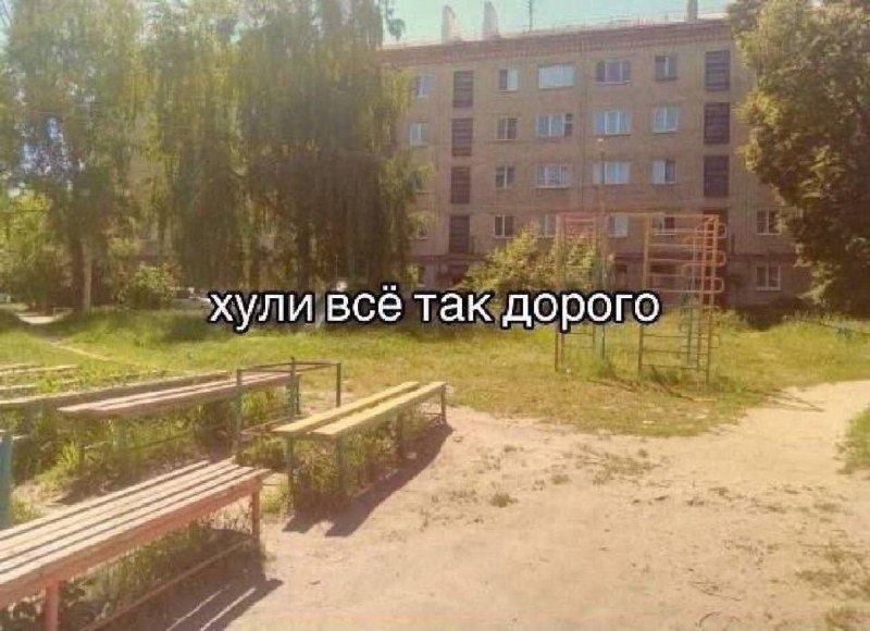 Двор со старыми скамейками и детской площадкой на фоне многоэтажки, сопровождаемый фразой «Хули всё так дорого».