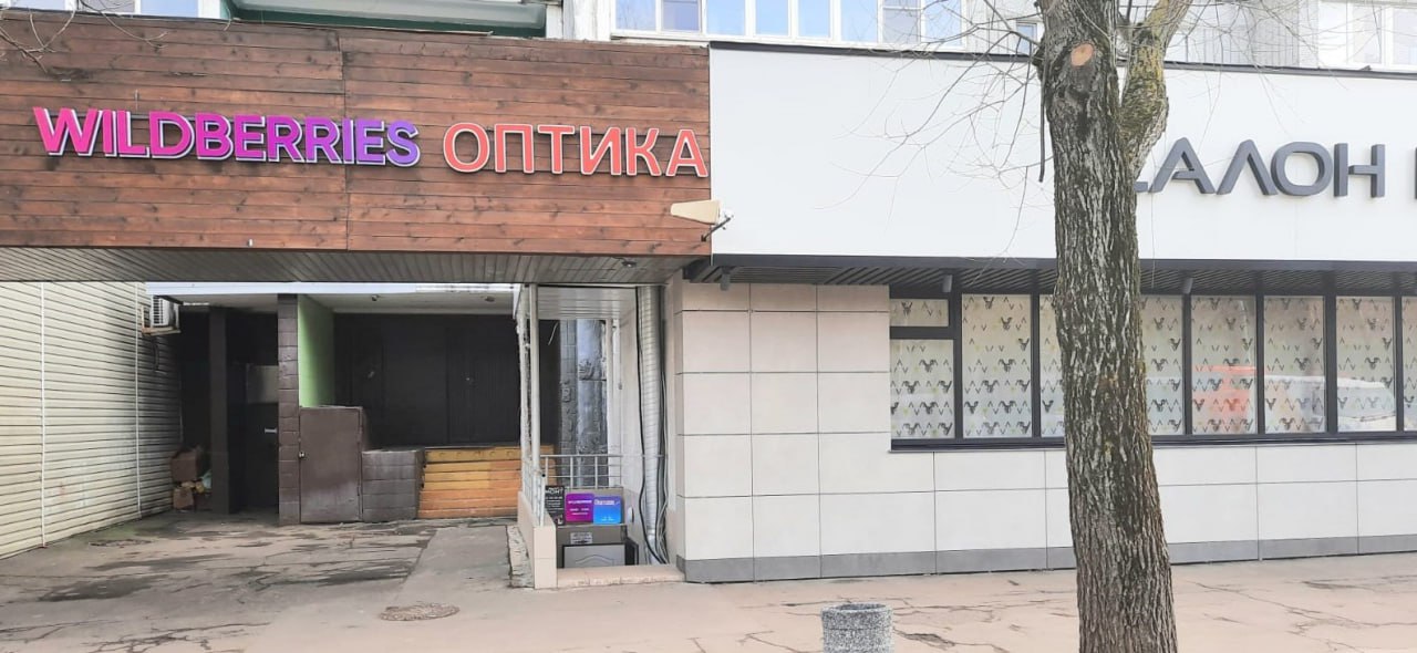 Wildberries оптика Вывеска "Wildberries оптика" на здании, часть улицы с лужами и деревом