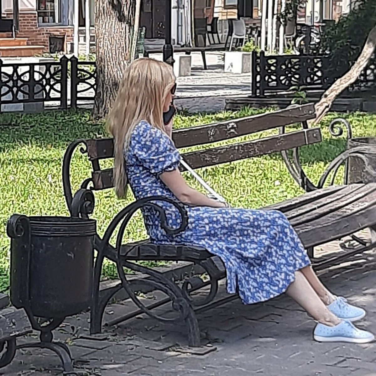 Женщина с тростью на скамейке Блондинка в синем платье сидит на скамейке с тростью и разговаривает по телефону.
