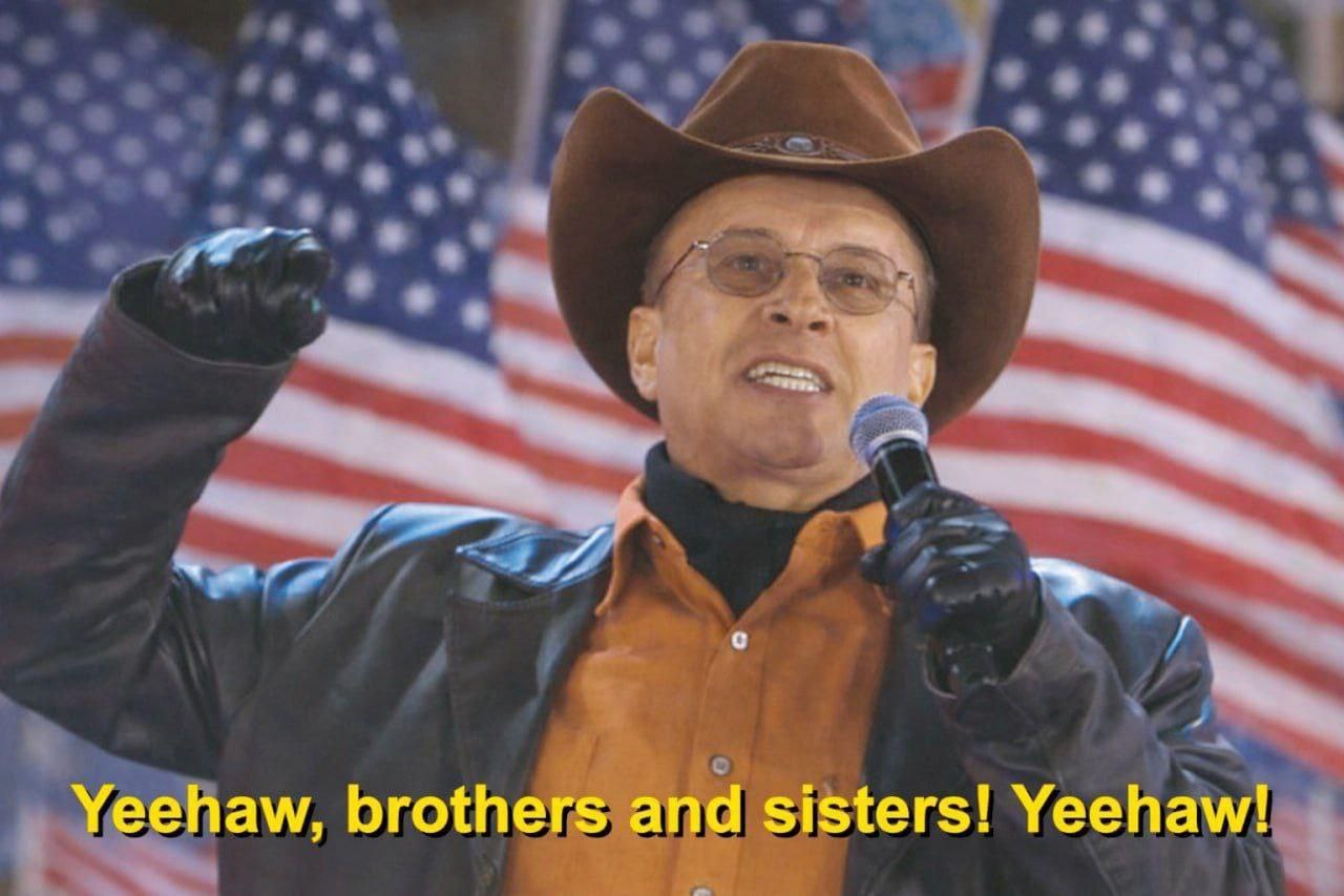 Мужчина в ковбойской шляпе, кричащий "Yeehaw, brothers and sisters! Yeehaw!" на фоне американских флагов.