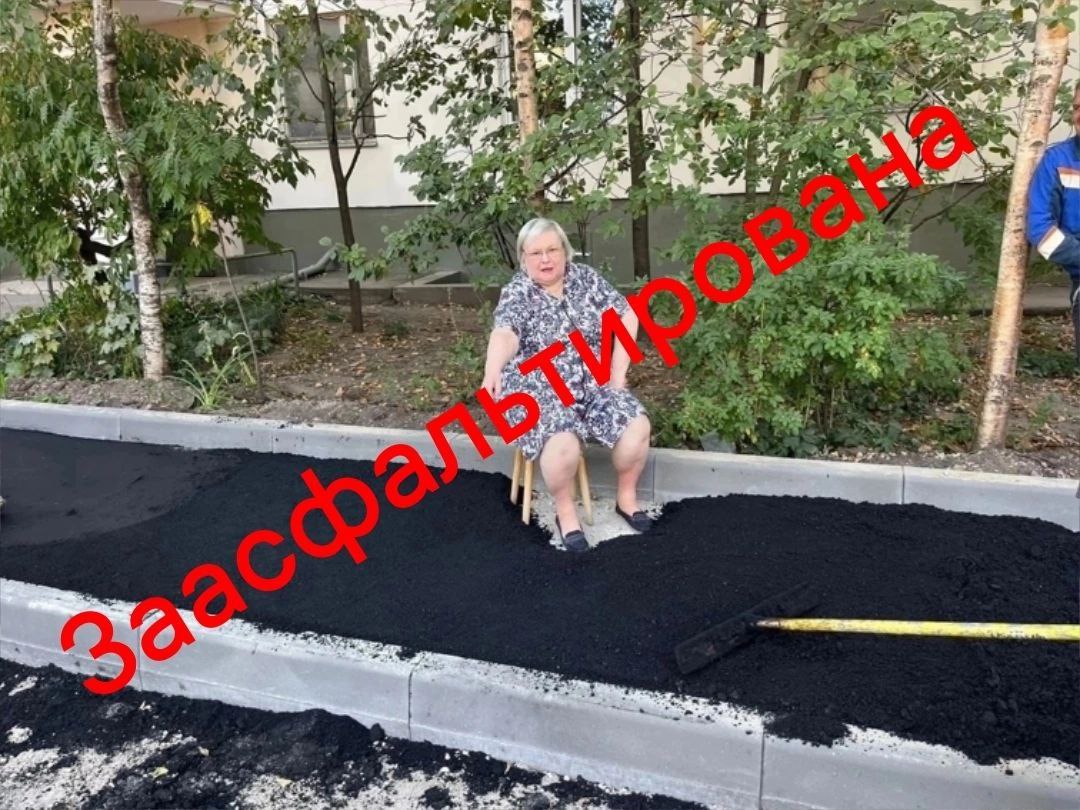 Заасфальтирована Пожилая женщина сидит на стуле посреди свежеуложенного асфальта на тротуаре, а рядом с ней лежит грабли.